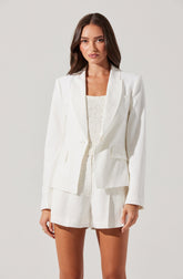 A woman is wearing a white tailored blazer with a single-button closure, structured shoulders, and flap pockets. She pairs it with matching high-waisted white shorts that have a loose fit and pleats. Underneath, she has on a cream-colored, textured sleeveless top.